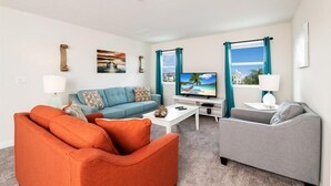 Een woonkamer met een blauwe bank, een oranje fauteuil, een grijze bank, een flatscreen televisie en een raam met teal gordijnen.