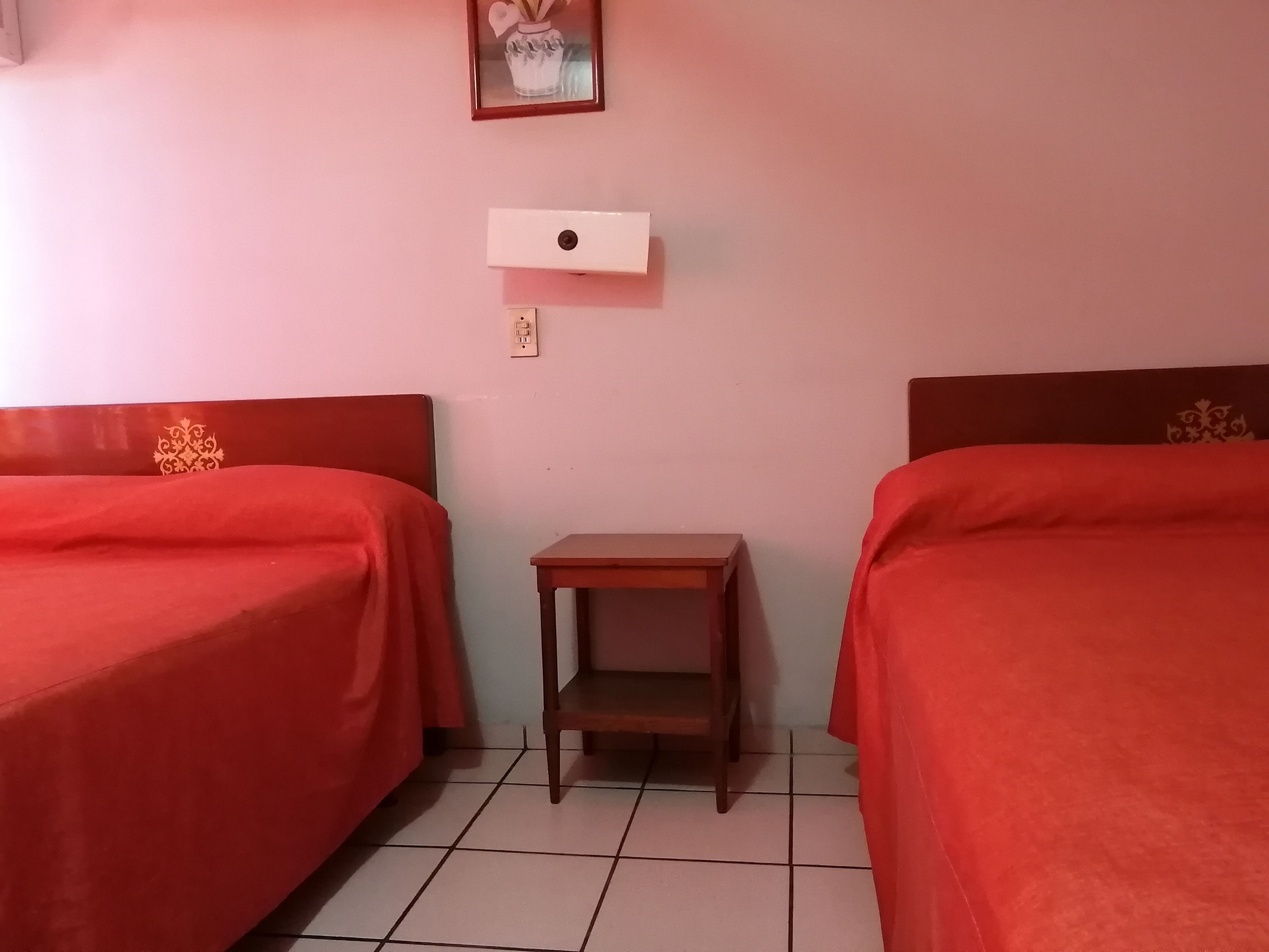 A room with two single beds, each with a red bedspread and a wooden headboard. There is a small wooden nightstand between the beds.