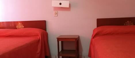 A room with two single beds, each with a red bedspread and a wooden headboard. There is a small wooden nightstand between the beds.
