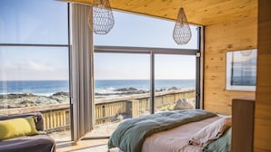 A bedroom with a large window overlooking the ocean, a bed with pillows, and a wooden ceiling. - Casas Punta Costanera (Pichilemu)