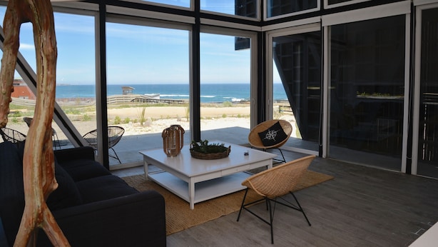 A modern interior with large windows, a white table, black sofa, and wicker chairs. - Casas Punta Costanera (Pichilemu)