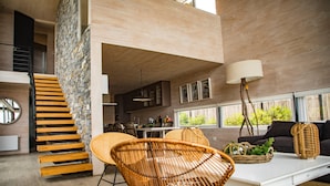 A modern living room with a staircase, wicker chairs, a white coffee table, and a large window with a view of greenery. - Casas Punta Costanera (Pichilemu)