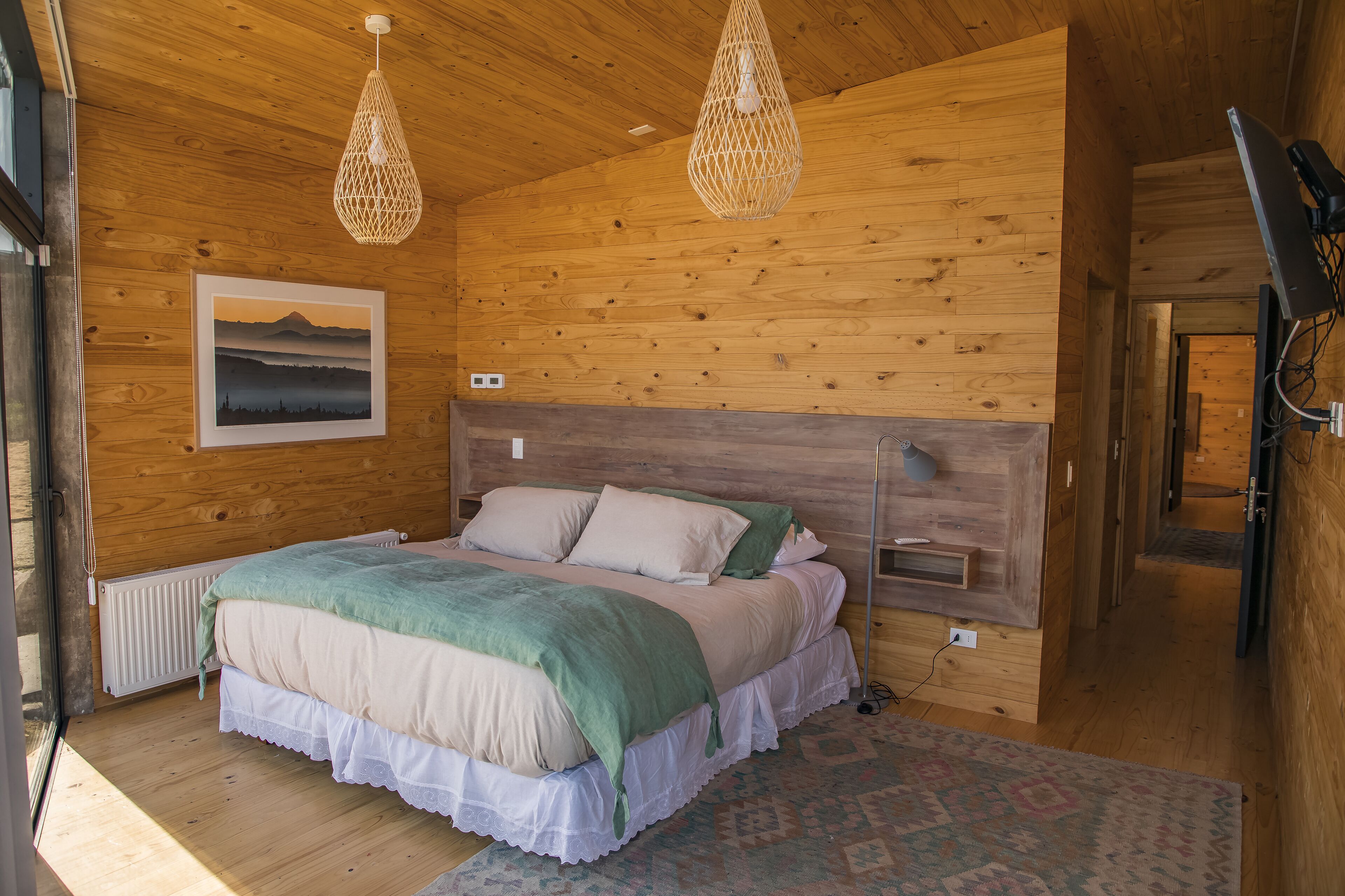 A wooden-paneled bedroom with a bed, two hanging lights, a framed picture on the wall, and a radiator.
