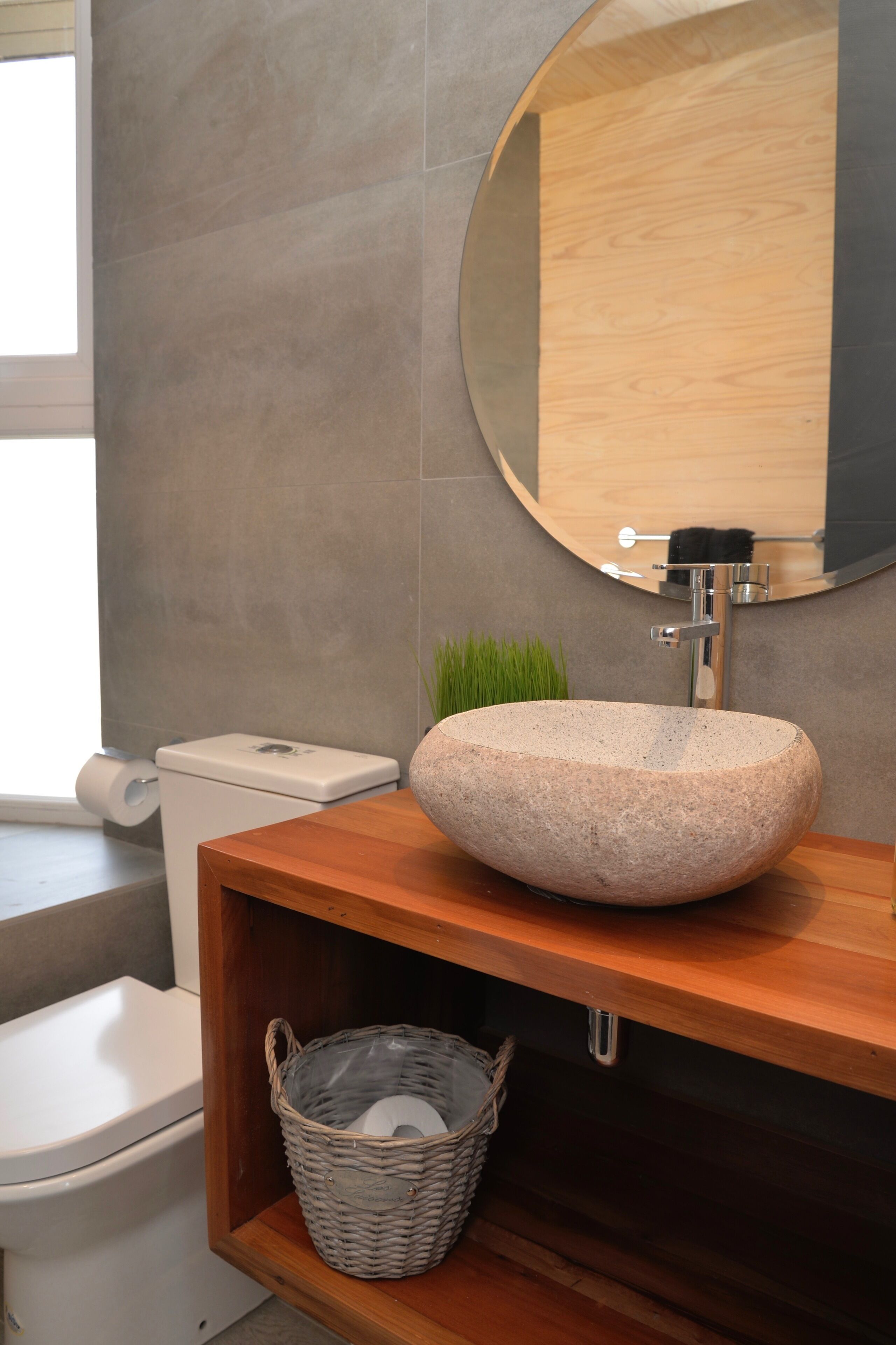 A bathroom with a stone sink, wooden vanity, a round mirror, a toilet, and a wicker basket.