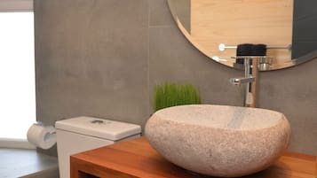 A bathroom with a stone sink, wooden vanity, a round mirror, a toilet, and a wicker basket.