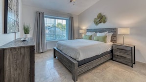 Un dormitorio con cama, mesitas de noche, ventilador de techo, ventana con cortinas, una planta y una obra de arte en la pared.