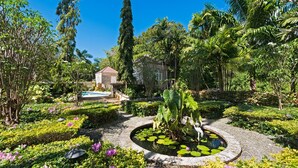 Un jardín con un estanque circular, diversas plantas y un edificio al fondo.