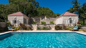 Una piscina rodeada de abundante vegetación, con dos edificios de piedra, cada uno con techo rojo y ventanas arqueadas.