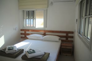 Una cama bien hecha con sábanas blancas, cabecera de madera y dos mesitas de noche con toallas dobladas.