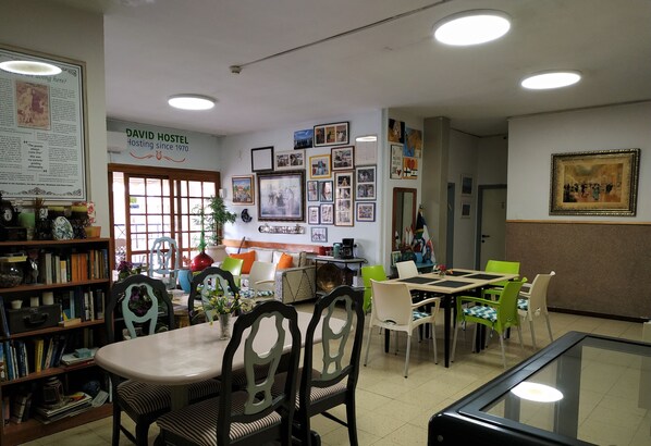 A cozy dining area with a variety of chairs and tables, a bookshelf filled with books and decorative items, and a wall adorned with framed photos and a sign reading "DAVID HOSTEL Hosting since 1970". - David Hostel - Hostel (Tiberias)