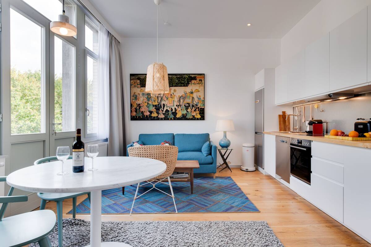 A modern kitchen with white cabinets, a dining area with a round table and chairs, and a living room with a blue sofa and a large painting.