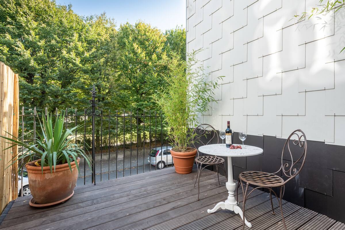 A wooden deck with a white table and metal chairs, surrounded by a black metal fence and a white wall with a geometric pattern.