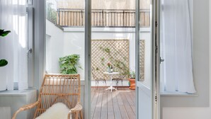 A room with a sliding glass door leading to an outdoor patio area. Inside, there is a wicker chair with a fluffy cushion, a small white table with a vase and a bottle, and a potted plant. - Europea Newton Boutique Hotel Residences (Brussels)