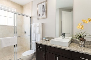 A modern bathroom with a glass shower enclosure, a white toilet, dark cabinets, a granite countertop, a white sink, a large mirror, and a vase with yellow flowers.