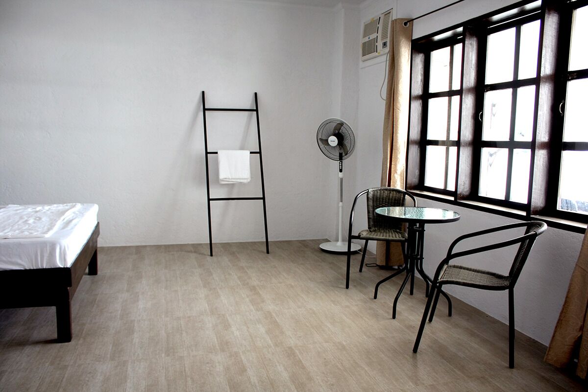 A room with a bed, a standing fan, a table with chairs, and a window with curtains.