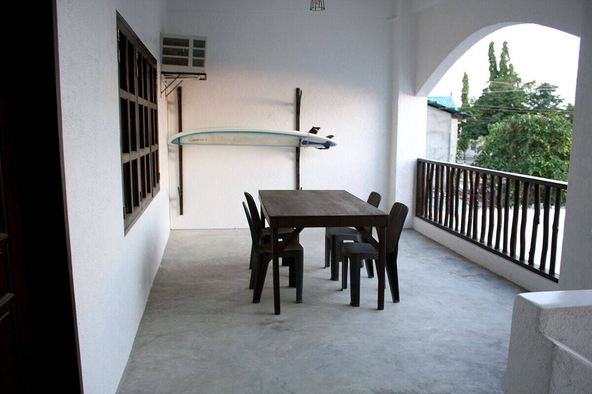 A balcony with a table and chairs, a surfboard, and a view of trees and a building.