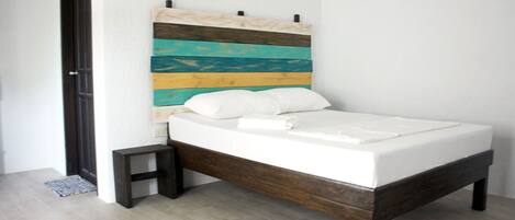 Una cama con estructura de madera y un cabecero colorido, una mesita de noche pequeña y una puerta a la izquierda.