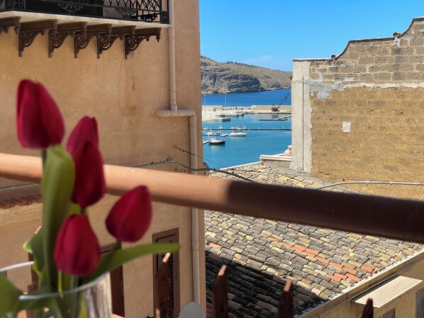 A balcony with a view of a harbor, a building with a stone wall, and a vase with red tulips. - Suite de la Sicilienne Deluxe in the center near the port and 200 mt from the sea (Castellammare del Golfo)
