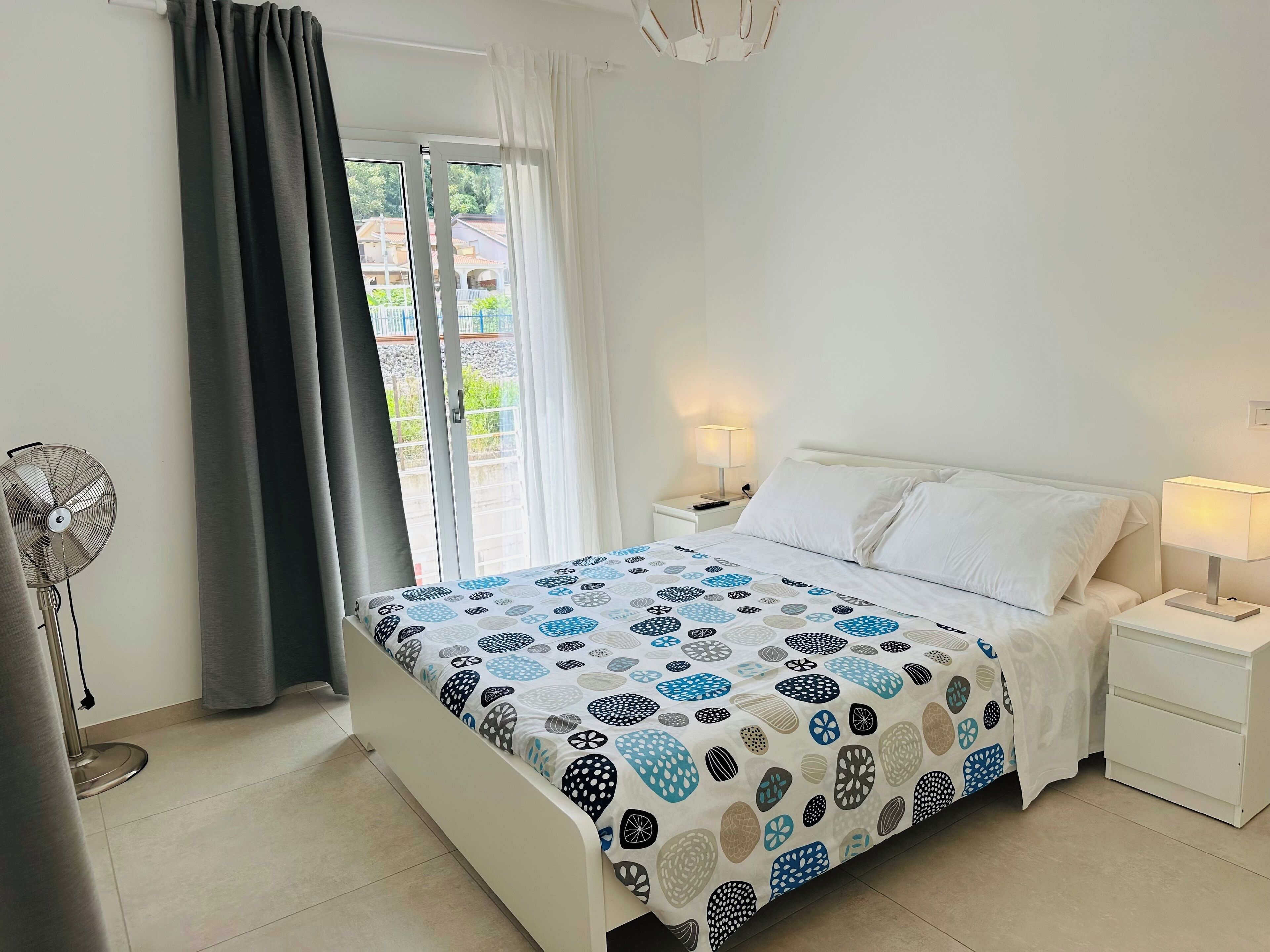 A bedroom with a bed, two bedside tables with lamps, a standing fan, and a window with curtains.