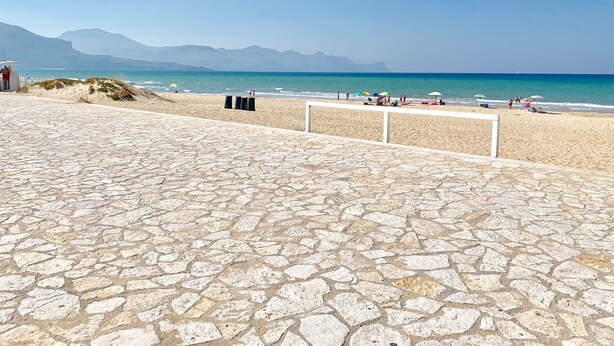 Una playa con un sendero de piedra que lleva a la orilla, algunas sombrillas y gente relajándose junto al agua.