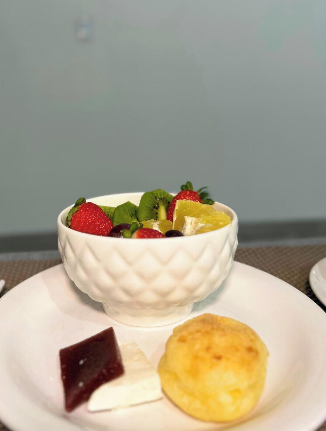 Un tazón blanco con ensalada de frutas sobre un plato blanco, acompañado de una galleta y un trozo de chocolate.