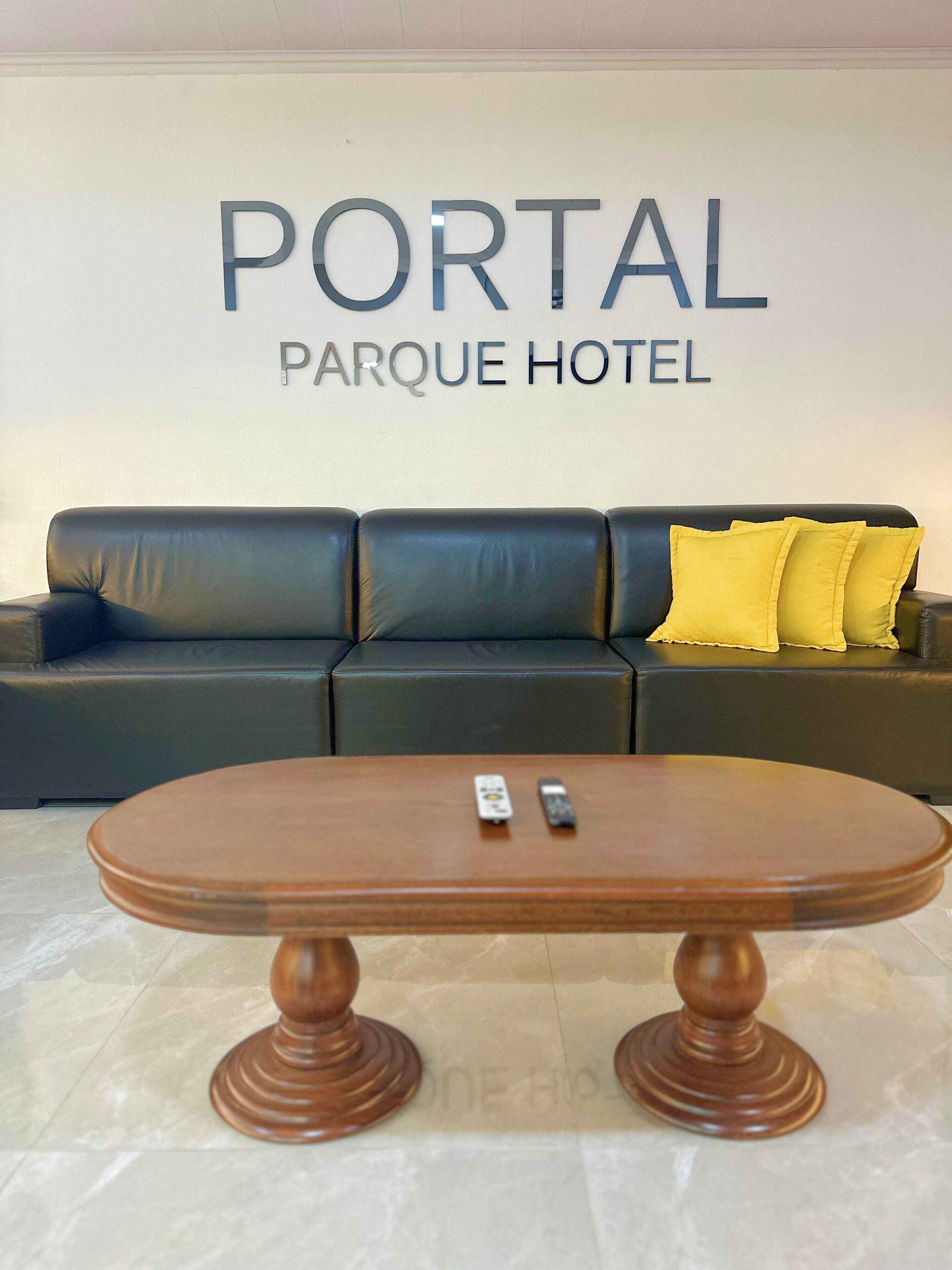 A reception area with a sign reading "PORTAL PARQUE HOTEL" above a black leather sofa with yellow pillows. In front is a wooden coffee table with two remotes on it.