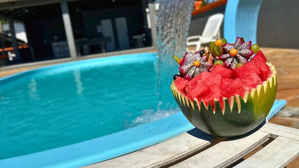 A bowl of watermelon and fruit on a poolside chair with a pool and a building in the background. - Portal Parque Hotel (Francisco Beltrao)