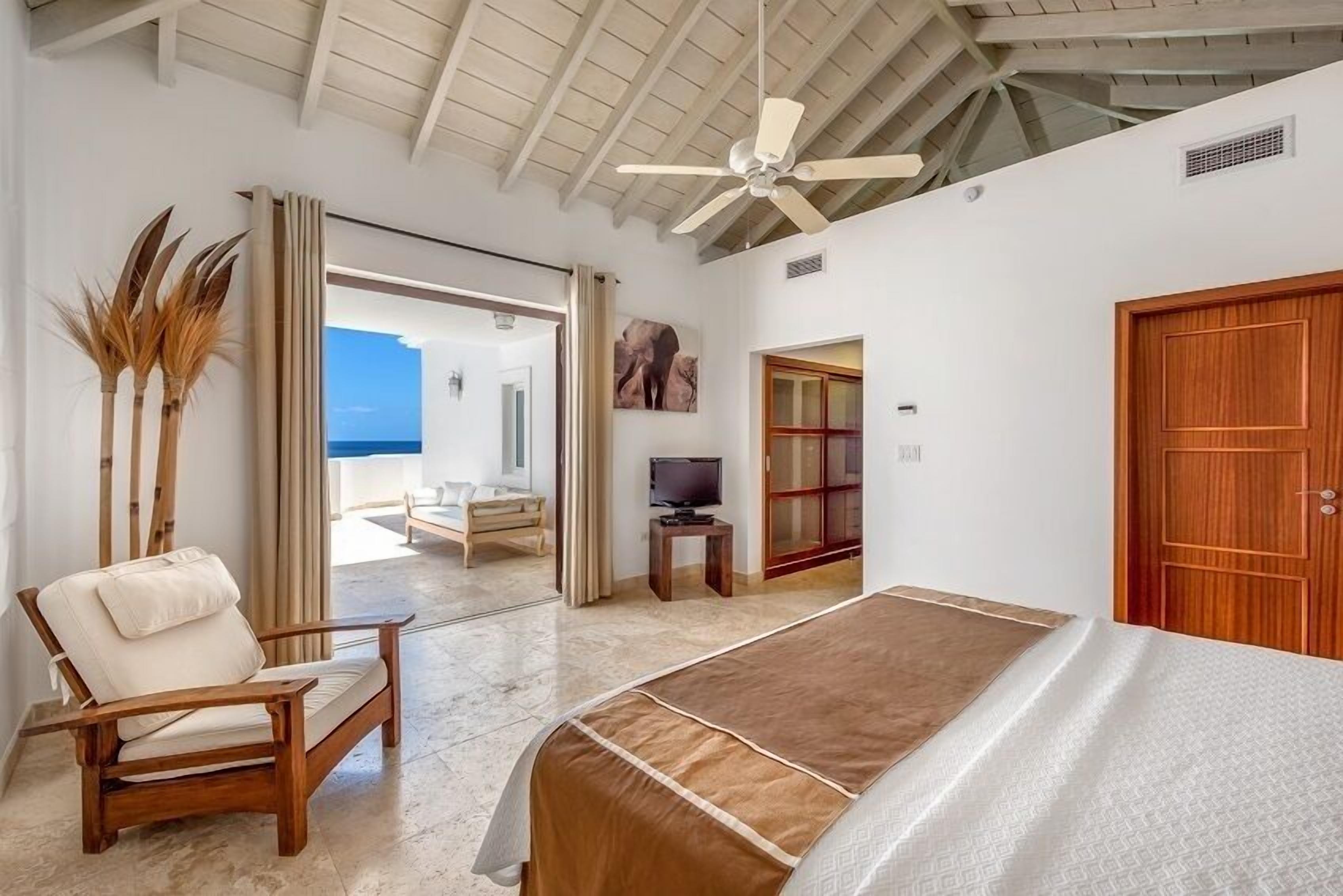 A bedroom with a bed, a wooden chair, a television, and a view of the ocean.