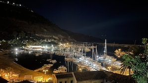 Un port de plaisance de nuit, avec des bateaux amarrés, des bâtiments à l’architecture traditionnelle et une colline en arrière-plan.