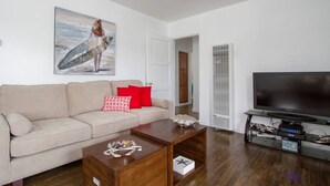 Ett vardagsrum med en beige soffa, röda och vita kuddar, ett soffbord i trä, en platt-TV och en tavla som föreställer en surfare.