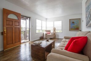 Una sala de estar con puerta de madera, sofá color beige y cojines rojos, una mesa de centro y una silla con cojín rojo.