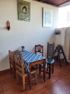Un comedor con una mesa cubierta por un mantel estampado, sillas de madera, un taburete de madera y una lámpara fijada a la pared.