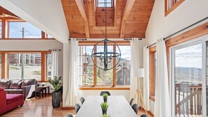 A spacious dining area with a long table, wooden ceiling beams, large windows, and a view of the surrounding landscape. - 34 chemin Blanc (CITQ: 280814) (Stoneham-et-Tewkesbury)