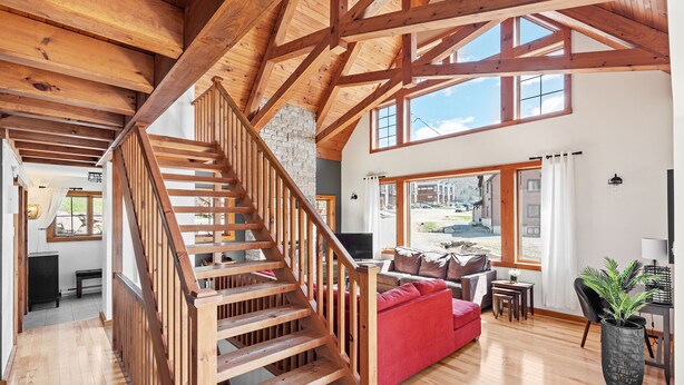A spacious living area with a wooden staircase, a red sofa, and large windows offering a view of the outside. - 34 chemin Blanc (CITQ: 280814) (Stoneham-et-Tewkesbury)