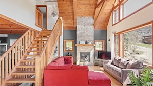 A spacious living room with a red sectional sofa, a grey sofa, a stone fireplace, wooden staircase, and large windows offering a view of the outdoors. - 34 chemin Blanc (CITQ: 280814) (Stoneham-et-Tewkesbury)