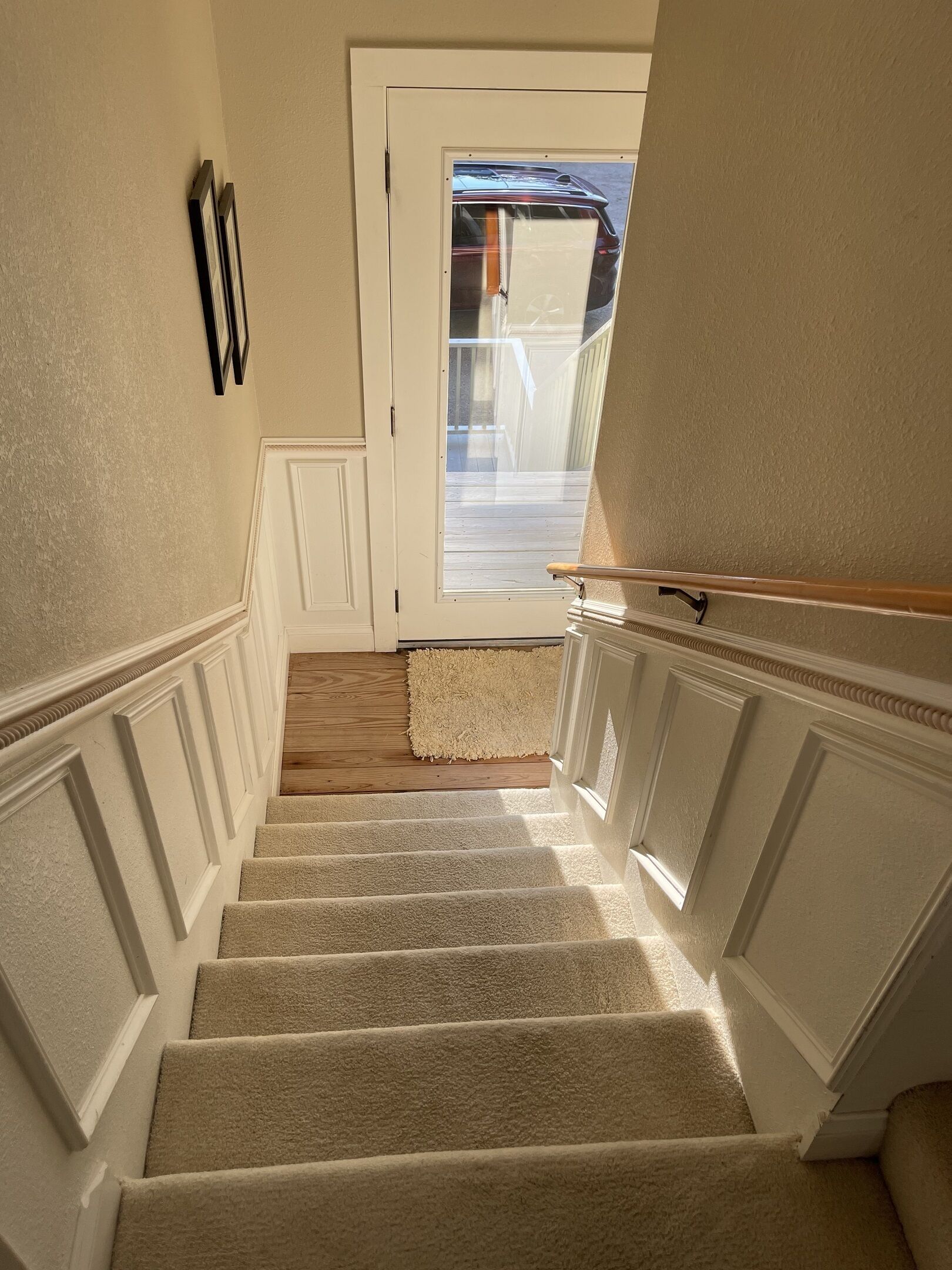 A carpeted staircase with a wooden floor at the top, white wainscoting, and a door leading outside.