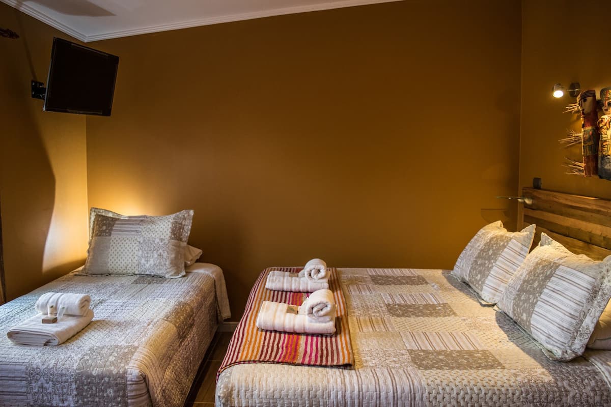 Ein Hotelzimmer mit zwei Betten, jeweils mit gemustertem Bettbezug und Kissen. An der Wand ist ein Flachbildfernseher angebracht, ein kleiner Tisch mit Handtüchern und eine Wandleuchte.