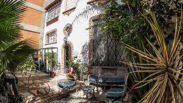 Un patio al aire libre con mobiliario de mimbre, plantas en macetas y un edificio blanco con detalles decorativos.
