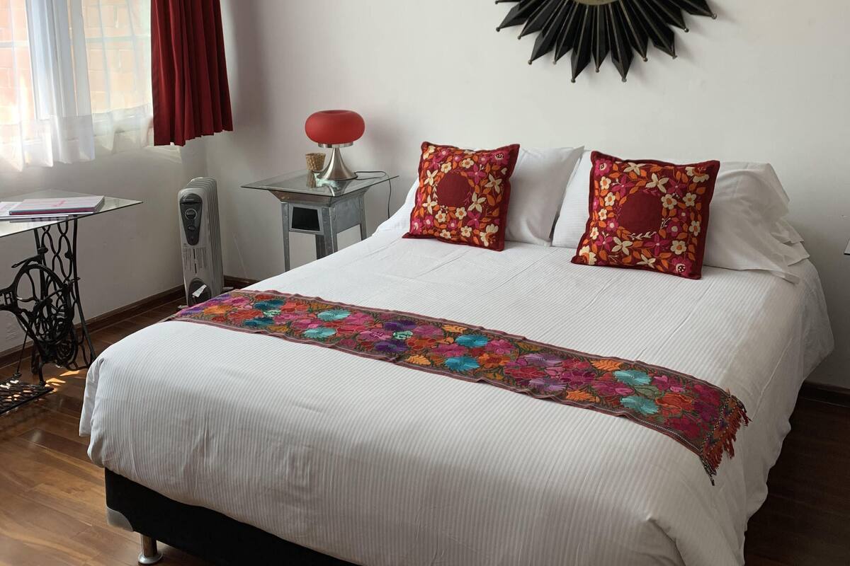 A neatly made bed with a floral bedspread, a red lamp, and a sunburst mirror on the wall.