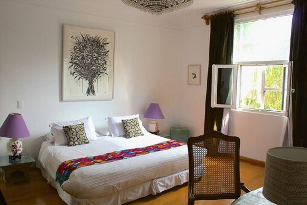 A bedroom with a bed, two bedside lamps, a chair, a window with curtains, and a wall-mounted artwork.