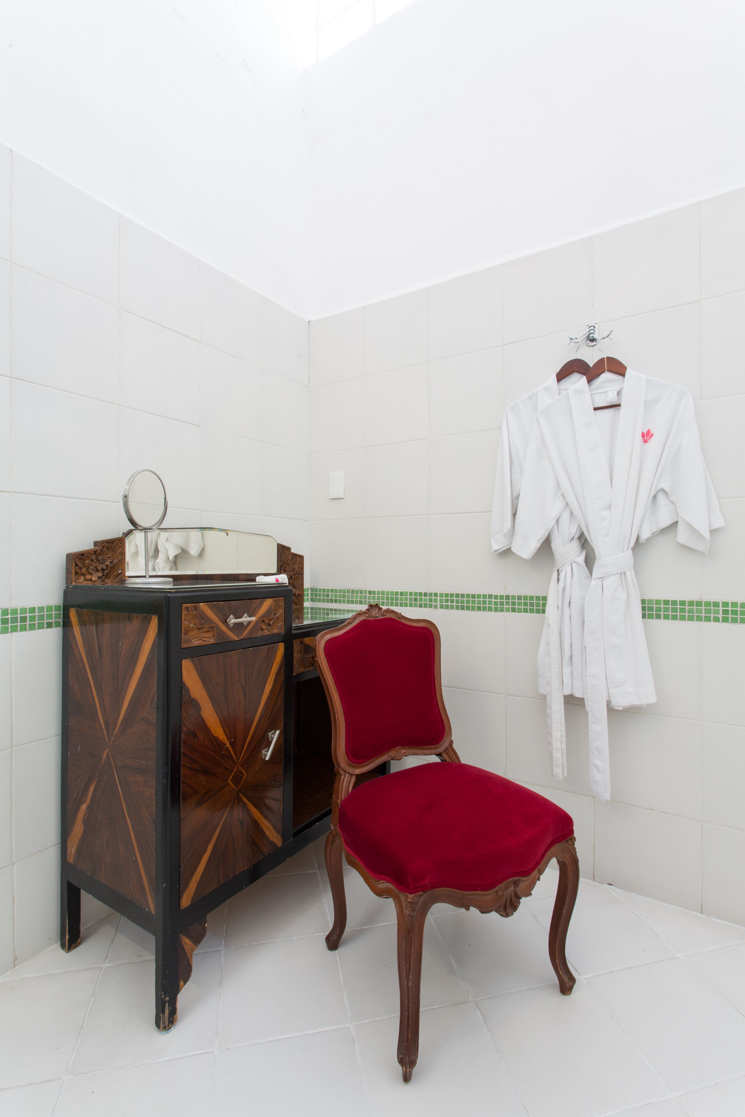 A bathroom with a vanity, a mirror, a red chair, and a white robe hanging on the wall.