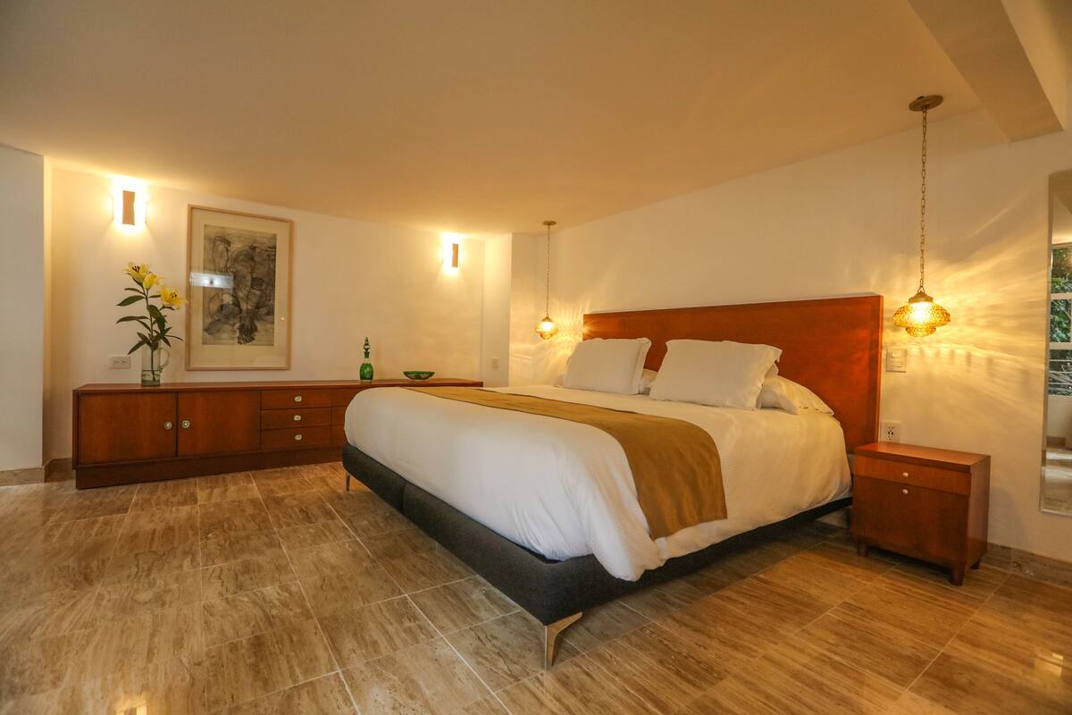 A spacious bedroom with a large bed, a wooden nightstand, a bedside table, a wall-mounted light, and a framed artwork on the wall.