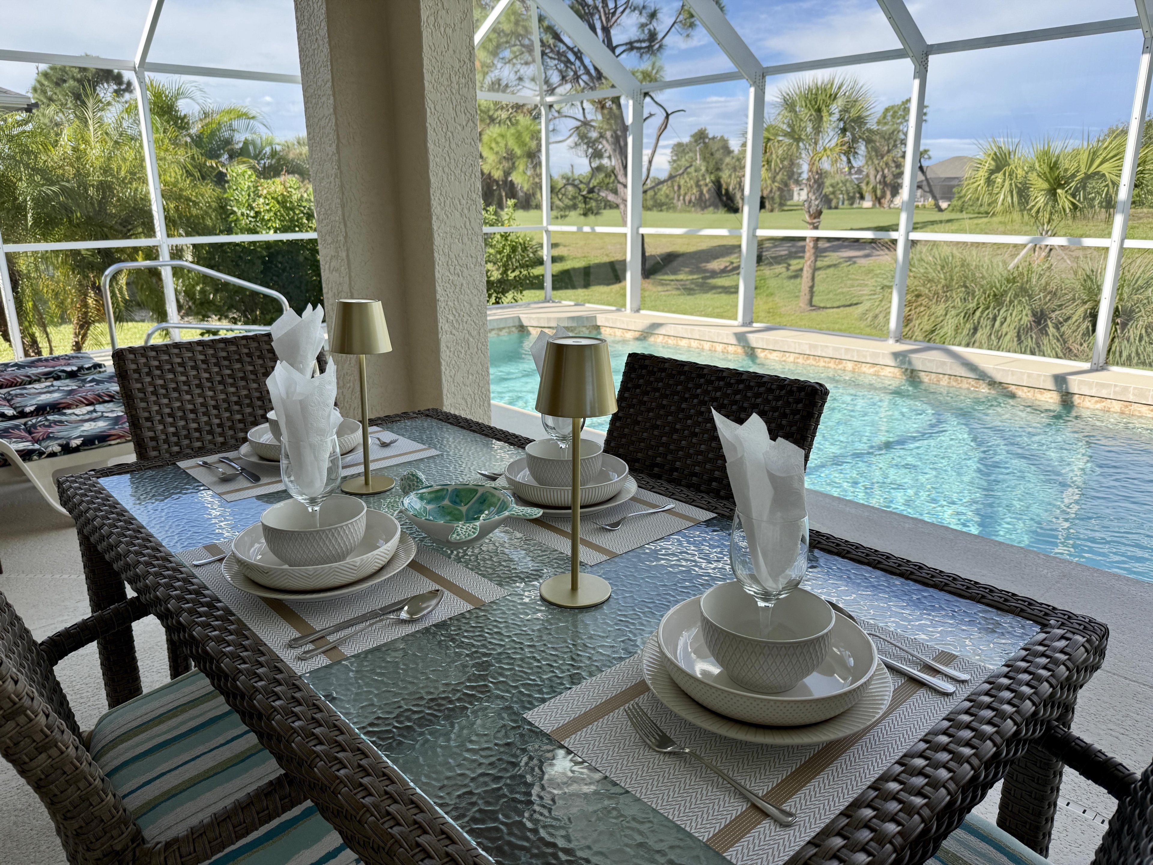 A glass-top table with white dinnerware and napkins, set against a backdrop of a pool and greenery.