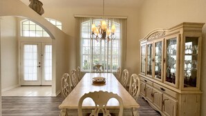 A dining room with a long wooden table, white chairs, a chandelier, and a glass-fronted cabinet.