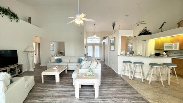 A spacious living room with a large sectional sofa, a coffee table, and a TV. There is a kitchen area with a bar and stools.