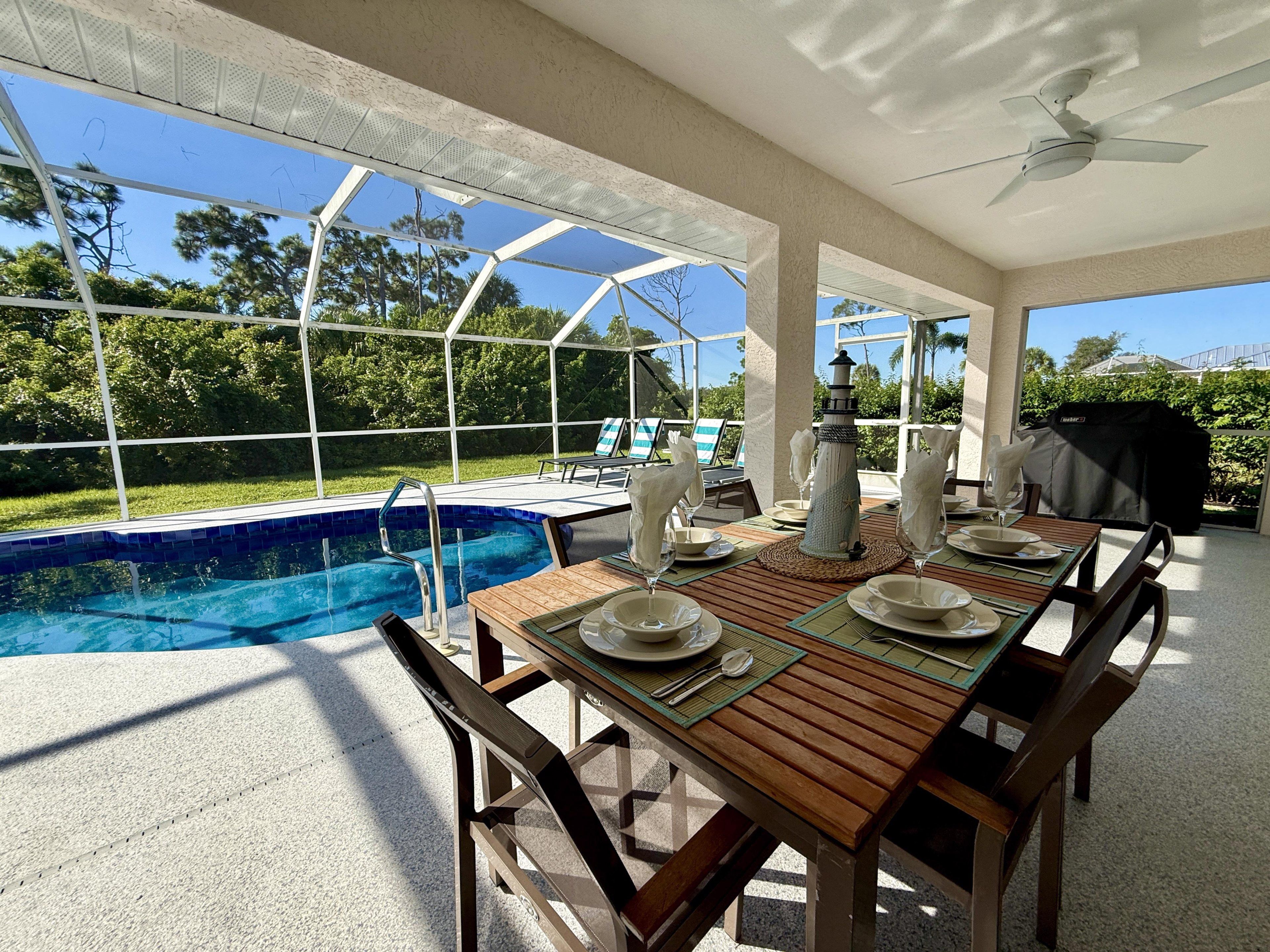 Un área de comedor al aire libre con piscina, mesa de madera, sillas y un ventilador de techo.
