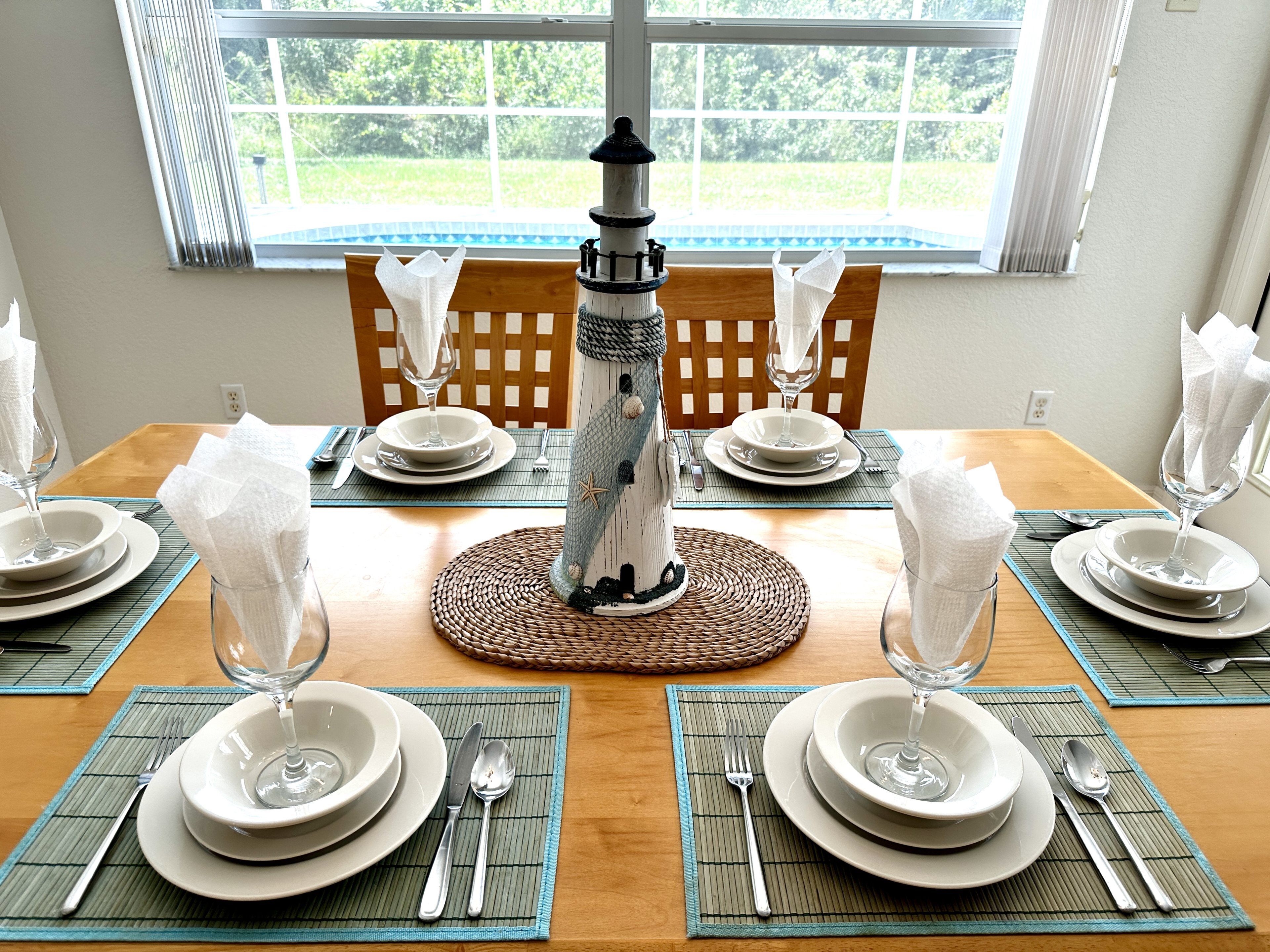 Mesa de comedor dispuesta para seis personas, con un centro de mesa decorativo de faro, sillas de madera y vistas a la vegetación exterior.