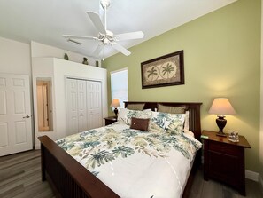 Une chambre à coucher avec un lit, un ventilateur de plafond, une table de chevet avec une lampe, un tableau encadré au mur et une porte donnant sur une autre pièce.