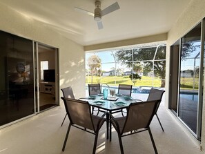 A dining area with a glass table and chairs, a ceiling fan, and a view of a swimming pool and greenery outside.