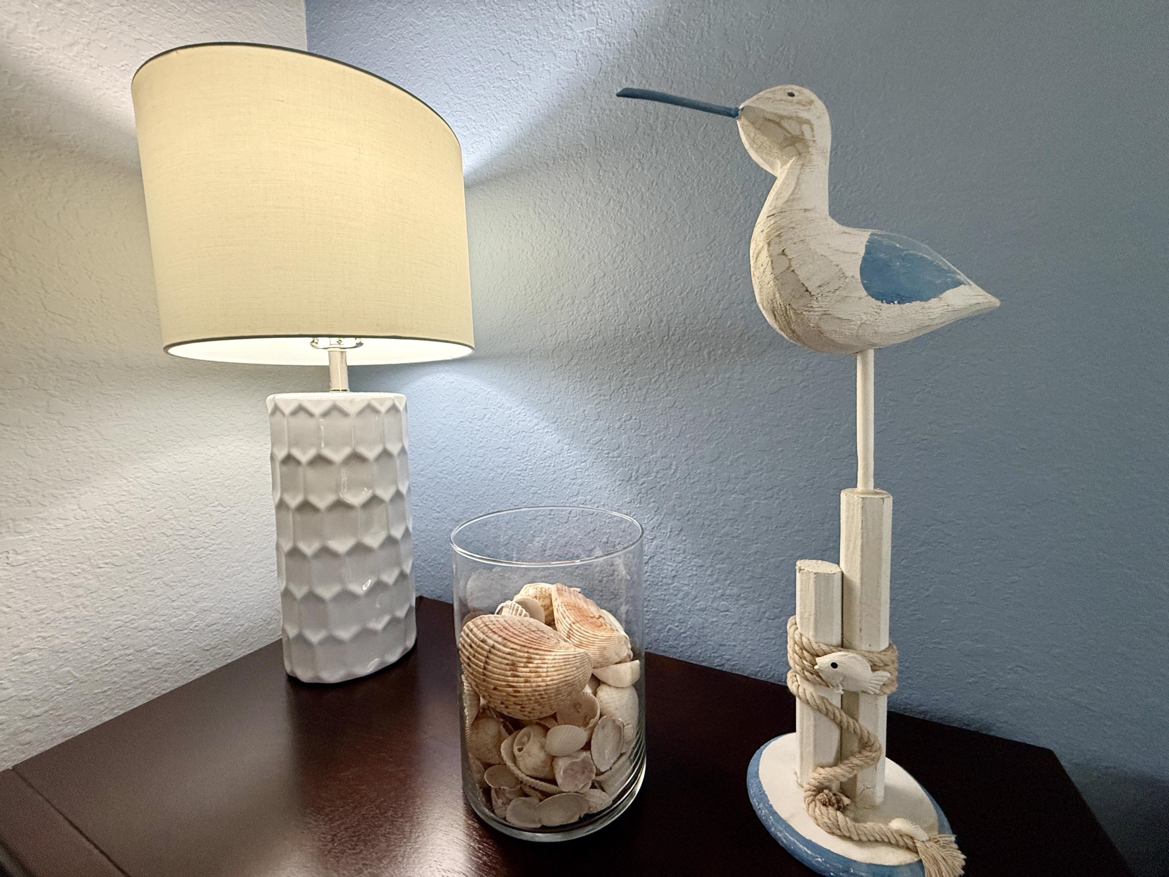 Een lamp, een glazen pot met schelpen en een vogelbeeld op een tafel.